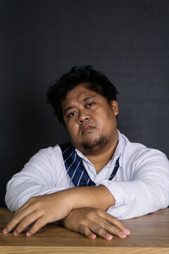 A highly unmotivated young Asian male corporate employee leans forward at a smooth wooden desk against a solid dark gray wall, resting both arms comfortably on the surface. The disheveled