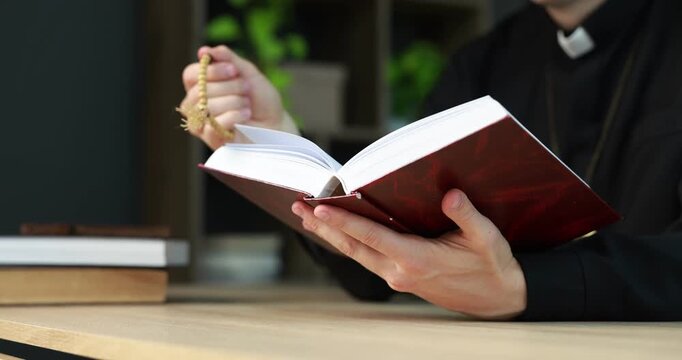 Priest in cassock with rosary reading Holy Bible at wooden table indoors, closeup