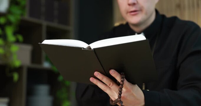 Priest in cassock with rosary reading Holy Bible indoors, closeup
