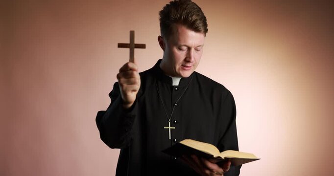 Priest in cassock with wooden cross reading Holy Bible on pale brown background