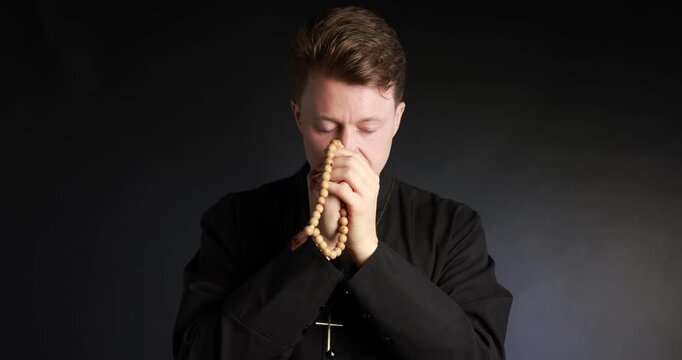 Priest in cassock with rosary praying on black background