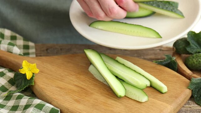 Woman putting pieces of cucumber from cutting board onto plate at wooden table, closeup