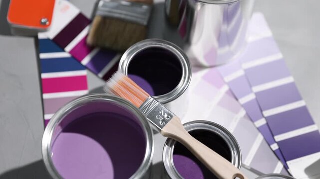 Cans with paint in shades of purple, brush and color palettes on light grey table, closeup. Camera moving in