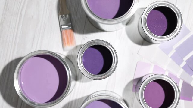 Cans with paint in shades of purple, brush and color palettes on white wooden table, flat lay. Camera moving in