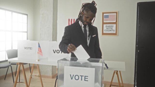 Man in suit places ballot into transparent ballot box beside vote booth and small us flags in voting building while smiling and gesturing with hand; civic pride.