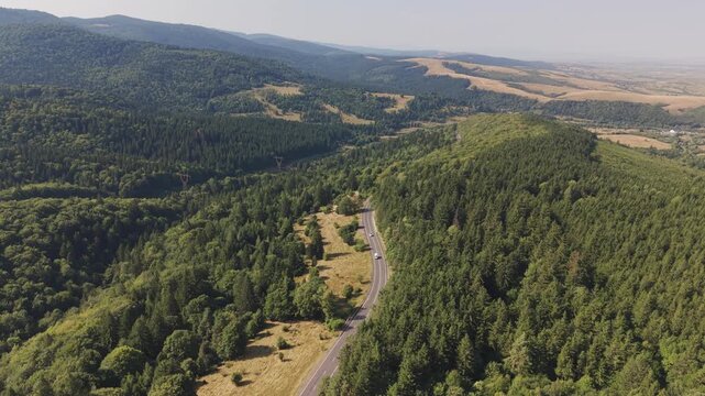 Drone flies backward above a winding mountain road in Romania, revealing vast forested hills, valleys and countryside stretching toward the horizon on a sunny summer day.