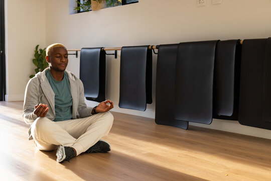 African American adult-male in hoodie meditating in yoga studio by barre with black mats