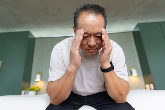 Senior Chinese man wearing white T-shirt sitting on bed edge pressing temples with black smartwatch