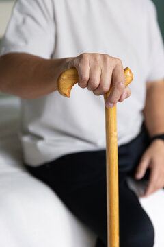 Senior Asian man sitting on light bed gripping wooden cane handle, wearing short-sleeve shirt
