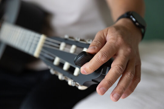 Senior Asian man tuning guitar headstock at home, holding clip-on tuner near nylon strings