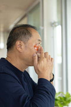 Senior Chinese man applying orange hydrogel patch beneath right eye in corridor wearing navy top