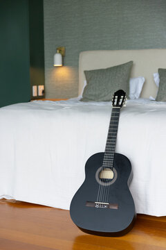 Black classical guitar is leaning against bed on warm wooden floor, green wall sconce is glowing