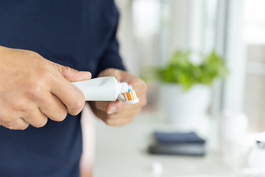 Senior Asian man squeezing toothpaste from tube onto orange white toothbrush at sink in navy top