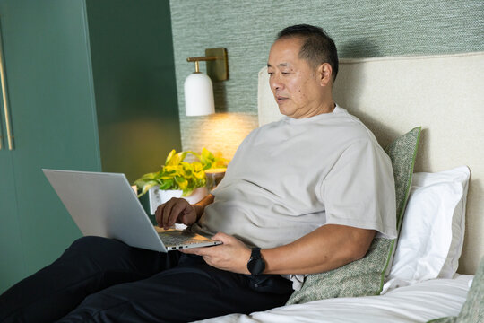Senior Chinese man working on silver laptop while leaning on green pillow in bedroom beside plant