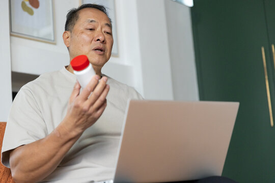 Senior Chinese man sitting on orange chair holding red-capped medicine bottle, using laptop at home