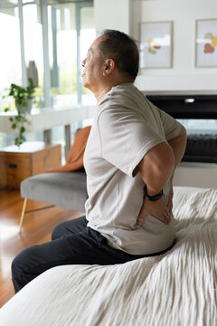 Senior Asian man sitting on bed at home holding lower back turning toward window wearing smartwatch
