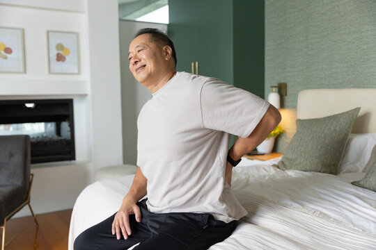 Senior Chinese man sitting on edge of bed, holding lower back, wearing beige T-shirt and smartwatch