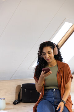 African American woman sitting on bench under skylight wearing headphones checking phone