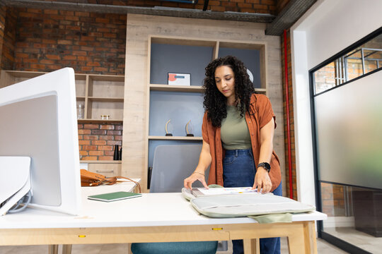 African woman standing at light-wood desk on right reviewing open portfolio with monitor