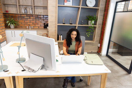 African woman wearing green top, rust cardigan working on laptop at office desk with large monitor