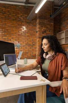 African American woman writing in notebook at desk in co-working space, pink headphones, copy space