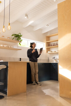African American man holding tablet and mug, standing at modern kitchen island under pendant bulbs