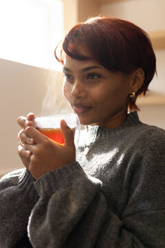 African American woman holding steaming mug of tea near bright window, wearing gray sweater