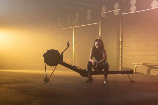 Woman sitting on rowing machine holding water bottle in gym wearing dark workout clothes