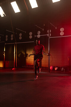 Male fitness trainee wearing red shirt jumping speed rope in dim gym with rig, kettlebells