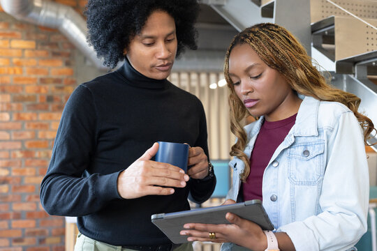 African American women colleagues in denim jacket reviewing tablet in folio office holding blue mug