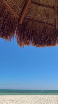 Vertical video of camera panning from shady thatched hut palapas to beautiful beach in Cancun Mexico.