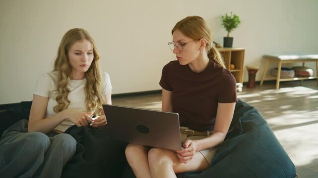 Two women meet for a psychology session while sitting on a beanbag in a light room with plants and furniture