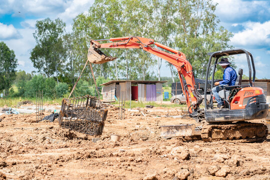 Mini excavator lifting a steel rebar cage frame for foundation footings at an outdoor construction site.