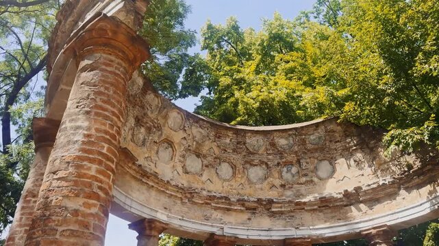 Low-angle close view of the weathered Tempietto d&rsquo;Arcadia in Parma&rsquo;s Parco Ducale, highlighting curved brick columns, faded plaster and decorative ruin details beneath summer trees.