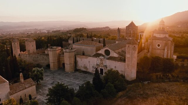 Cinematic aerial view of the historic Santa Maria de Poblet Monastery illuminated by warm golden-hour sunlight in Catalonia, Spain. The drone reveals the medieval monastery complex, towers, church and