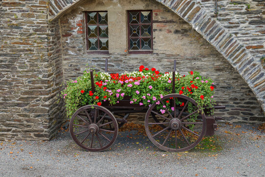 Vintage wooden cart decorated with blooming red and pink flowers parked against an ancient stone wall with a rustic window and architectural archway