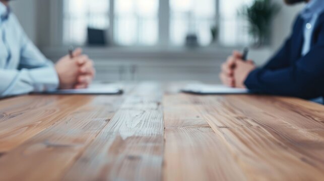 Blurred background of an office reveals two individuals in a professional meeting seated across a rustic wooden table with papers and pens
