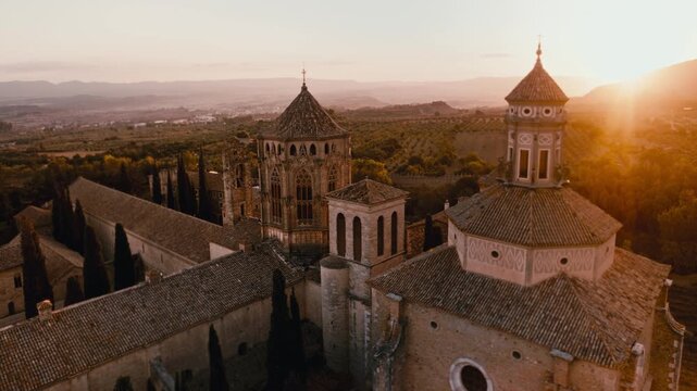 Close aerial drone view of Santa Maria de Poblet Monastery at golden hour, featuring its historic bell tower, tiled roofs and warm sunset light over the Catalan countryside.