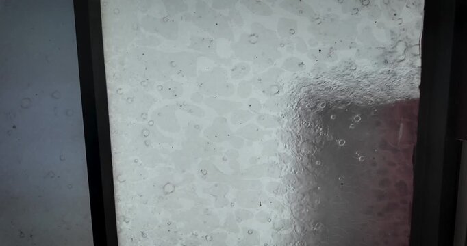 Heavy rainfall covers a window with moving droplets and flowing water while the urban landscape of Mexico City appears blurred in the background during a storm.