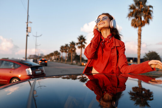 Smiling woman in red jacket listening to music with headphones on car roof. Concept of lifestyle, travel, and freedom. Warm sunset lighting creates mood.