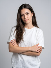 Fototapeta premium Portrait of a young woman with long dark hair wearing a white tshirt posing with arms crossed against a plain background.