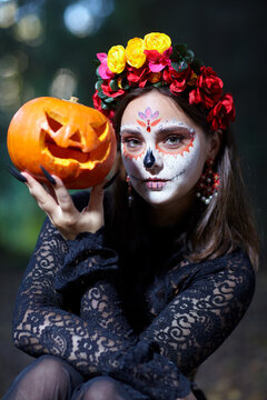A young woman dressed as Santa Muerte sits in a forest holding a Halloween pumpkin in hand.
