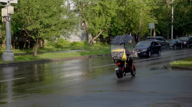 Food delivery courier on electric moped with rain canopy riding on wet city street during rainy weather