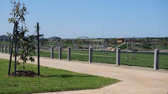 A newly developed suburban park in an outer&nbsp;suburb with footpath, immature staked saplings offering minimal shade, and a children's playground at an unshaded open green field. Melbourne Australia