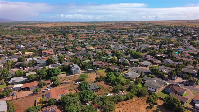 Aerial view over Kihei on Maui Hawaii island showing dense residential neighborhood with red tile roofs solar panels tropical trees quiet streets and golden dry fields under blue sky.
