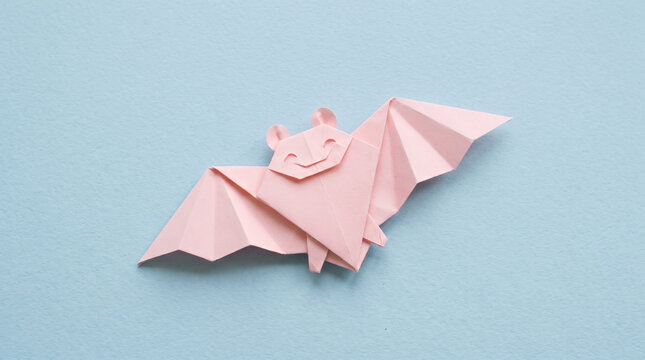 Cheerful round eared pink origami bat character on powder blue, cute halloween paper craft background for greeting cards