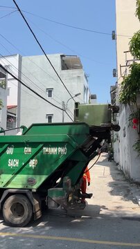 A green garbage truck is parked on a narrow street. The truck is partially elevated, with buildings and power lines in the background. Bright sunlight illuminates the scene.