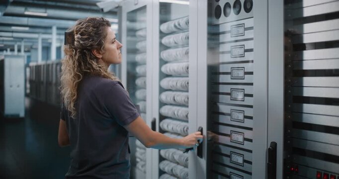 IT Specialist Holds Laptop Computer to Inspect Server Racks and Configure Cloud Infrastructure in Data Center Server Room. System Diagnostics and Hardware Maintenance Ensure Seamless Network Uptime.