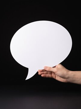 Hand holding a blank white speech bubble cutout against a black background for communication and message concepts.