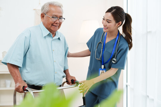 Young friendly female caregiver nurse in blue scrubs is smiling and helping Asian senior man practice walking with walker in cozy living room at nursing home. Physical therapy, healthcare concept